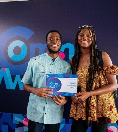 Two people smiling, holding an award certificate in front of a promotional backdrop at an event.