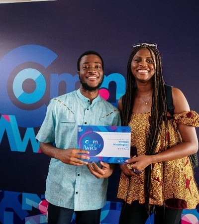 Two people smiling, holding an award certificate in front of a promotional backdrop at an event.