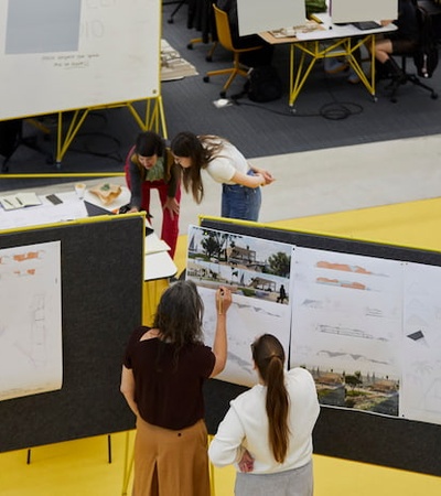 Three people are discussing over architectural drawings displayed on panels in a busy workshop environment.