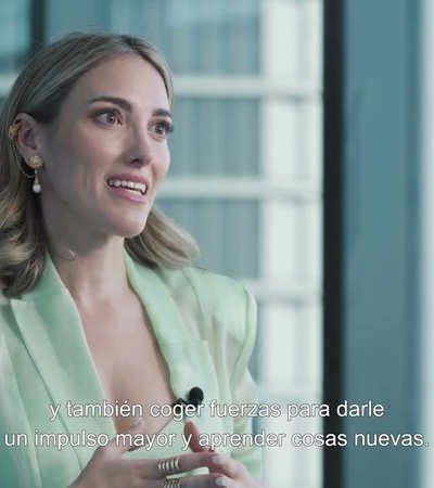 A woman gesturing while speaking at a professional event, with a background of a blurred window and subtitles on the screen.