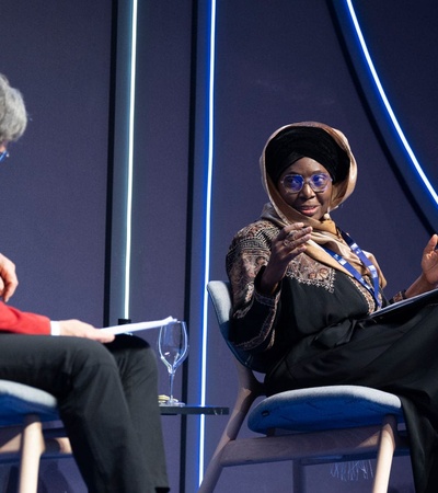 Two women engaged in a discussion on stage at a conference, one sitting on the left wearing a red jacket and the other on the right wearing a hijab and black dress.