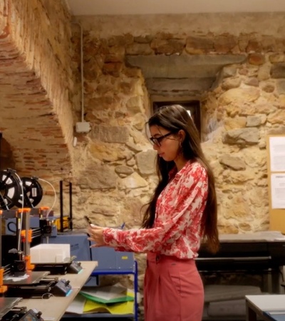 A woman is standing in a workshop with 3D printers and other equipment around her.