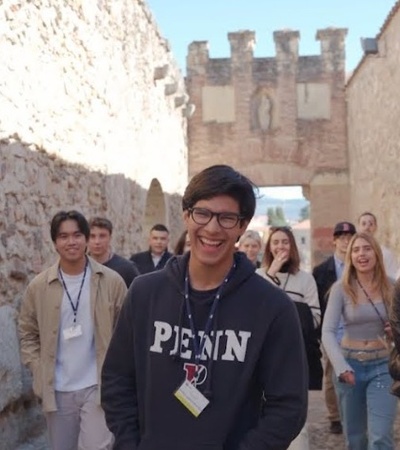 A group of young people walking along a historical stone-walled pathway, with a man in the foreground wearing a 'PENN' hoodie smiling at the camera.