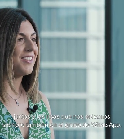 A woman with long brown hair in a green patterned top smiles while standing in a modern office environment.