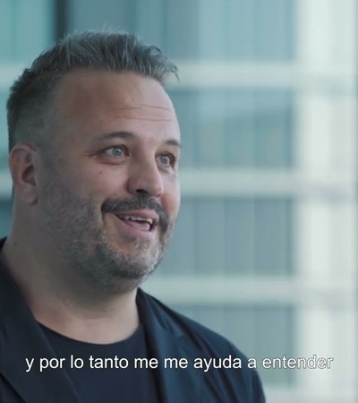 A man with a beard is smiling and speaking in a room with a modern interior and large windows in the background.