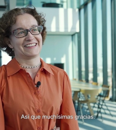 A smiling woman in an orange blouse stands in a modern office corridor with glass walls and blinds.