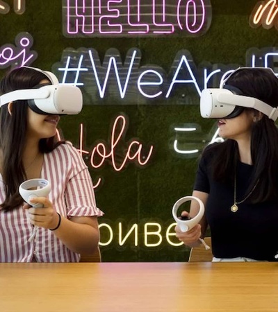 Two women wearing virtual reality headsets are interacting with each other in a room with a colorful neon sign wall in multiple languages.