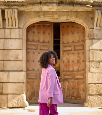 A woman in a pink suit smiles while walking past an old wooden door of a stone building.