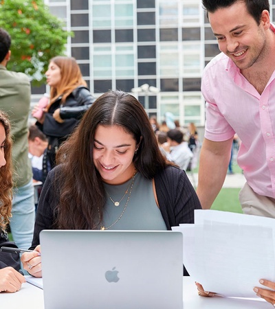 Four young adults are happily working on a project outdoors on a campus, using a laptop and papers.