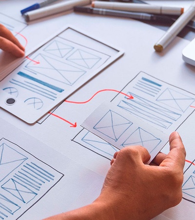 A person working on website wireframe sketches on a desk with a laptop and colored markers.