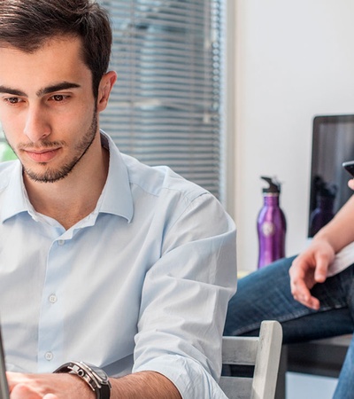 Two men in a modern office setting, one focused on a laptop and the other using a smartphone.