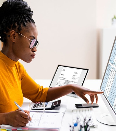 A woman working intently at her desk with two computer screens, one displaying spreadsheets.