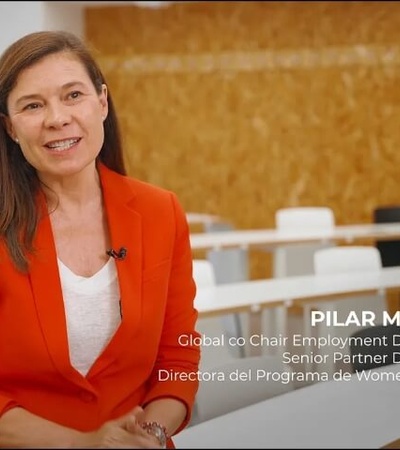 A woman in a red blazer is speaking in a classroom setting.