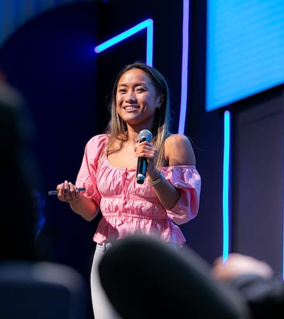 A woman in a pink blouse is speaking at a conference while holding a microphone.
