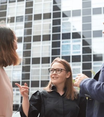 A group of three professionals are engaged in a conversation outdoors near a modern building.
