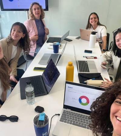 A group of six smiling women seated around a meeting table with laptops and a presentation visible.