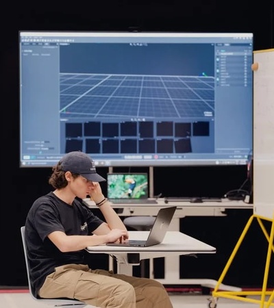 A young man is sitting at a table working on a laptop with a large screen displaying a digital interface in the background.