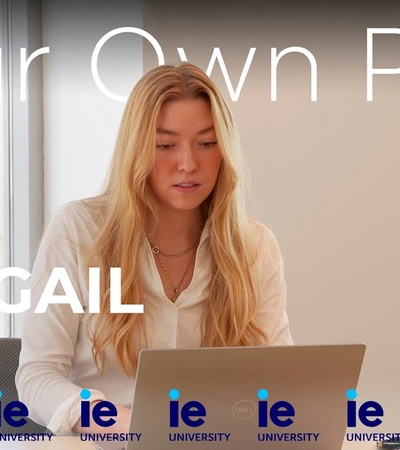 A young woman working on a laptop in an office setting, with promotional text for 'Your Own Path With ABIGAIL' at IE University.