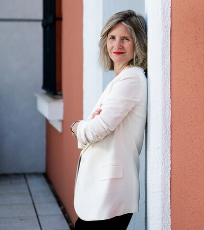 A woman in a white blazer leans against a wall, exuding confidence.