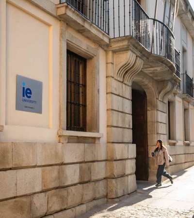 A woman is walking past the IE University building with a clear sky in the background.