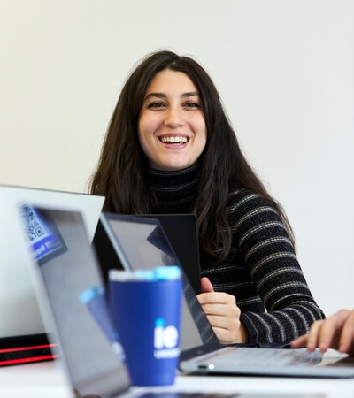 A smiling woman sitting at a table with laptops and a coffee cup.
