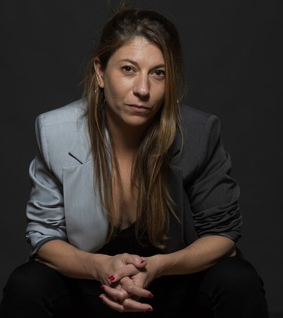 A woman sitting with a serious expression, wearing a blazer and looking directly at the camera.
