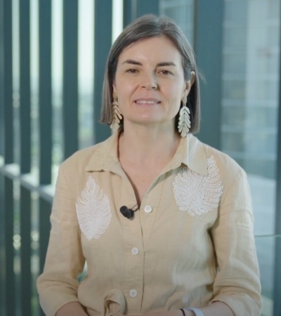 A woman with medium-length hair wearing a beige shirt with white embroidery stands in a building with a modern glass backdrop.