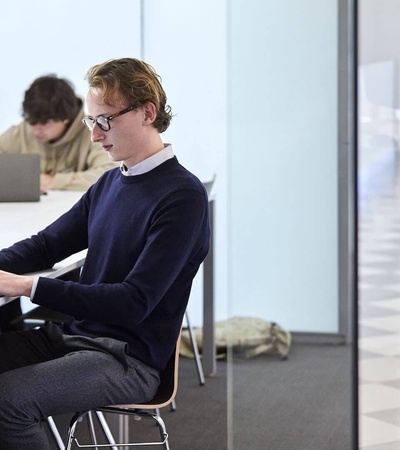A young man is using a laptop in a modern office environment with other people working in the background.