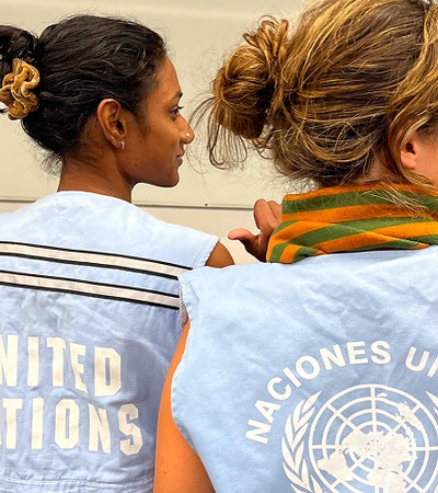 Two women are seen from behind, wearing blue vests with the United Nations logo.