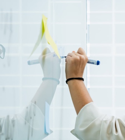 A woman is writing on a clear glass board with a marker while wearing glasses.