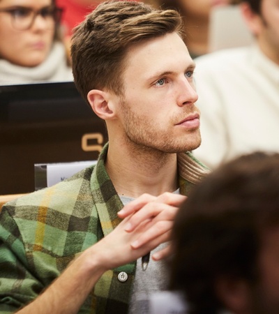 A young man with a green plaid shirt is listening attentively in a classroom setting.