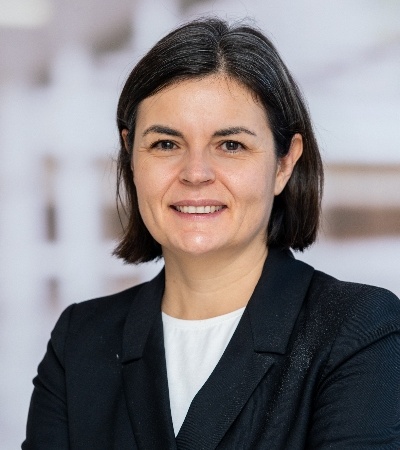 A professional portrait of a smiling woman with short, dark hair, wearing a black blazer over a white top.