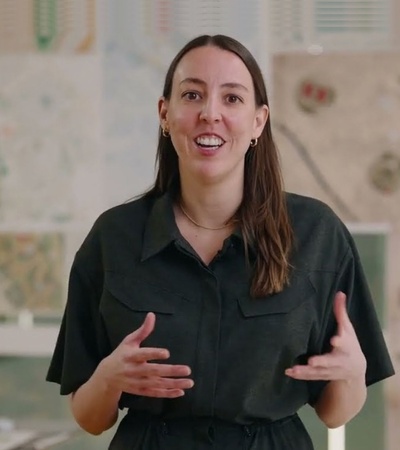 A woman stands in front of various maps and designs, engaging with the camera.