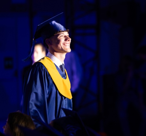A smiling graduate in a blue cap and gown stands proudly under stage lights.