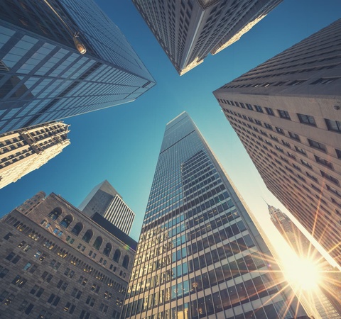 Looking up at towering skyscrapers against a clear blue sky with sunlight shining through.