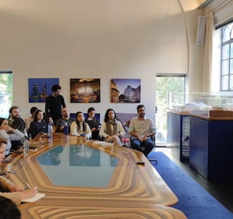 A man presenting to a group of people sitting around an oval table in a well-lit room with large windows and promotional posters on the walls.