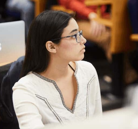 A young woman wearing glasses and a white top sits attentively in a classroom with a laptop in front of her.