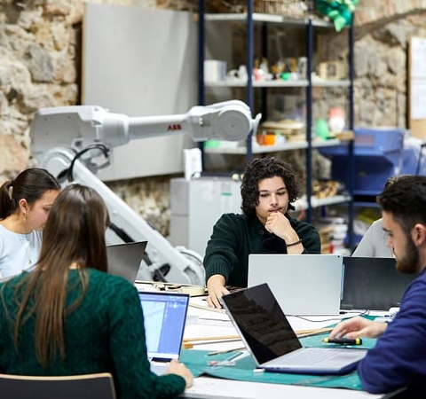 A group of students collaborating on laptops in a colorful workspace with a robotic arm in the background.