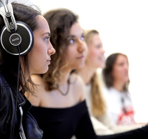 A young woman wearing headphones is sitting at a microphone in a studio, with other women in the background.