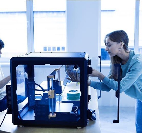 A woman operates a 3D printer while a man works at a nearby table in a bright room.