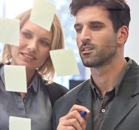 Two people writing on some post-it notes on a transparent blackboard.