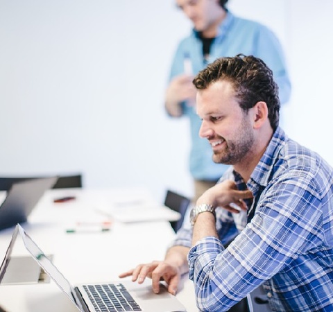 A man in a plaid shirt is working on a laptop at a conference table while another person stands behind him.