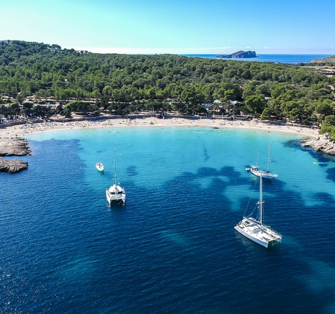 A serene beach with clear blue water and sailing boats anchored nearby, surrounded by lush greenery.
