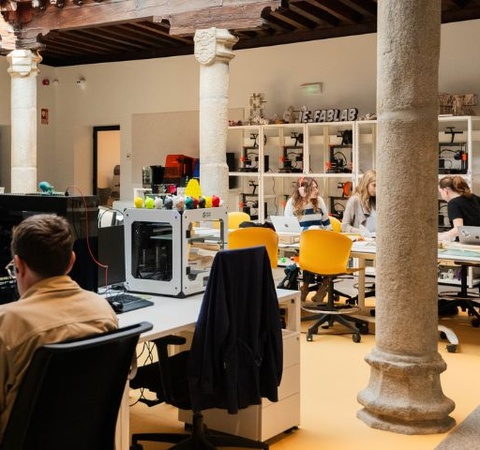 A modern coworking space with people working at desks and a 3D printer in the background.