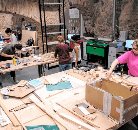 People are working collaboratively in a spacious workshop with various tools and materials on their desks.