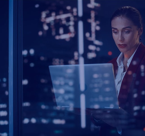 A woman in professional attire looking at a laptop screen while illuminated by city lights at night.