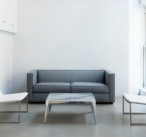 A minimalistic living room with a gray sofa, two white chairs, and a marble table against a backdrop of white walls and large windows.