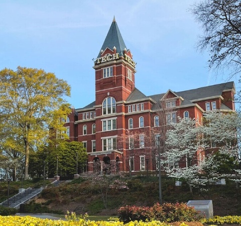 A historic brick building with a tower, surrounded by trees and flowering plants in a park-like setting.
