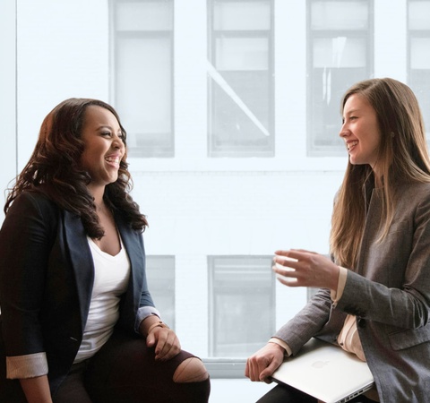 Two women are engaged in a lively conversation by a large window.
