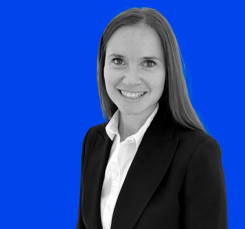 A smiling woman with a black blazer and white shirt stands against a blue background.